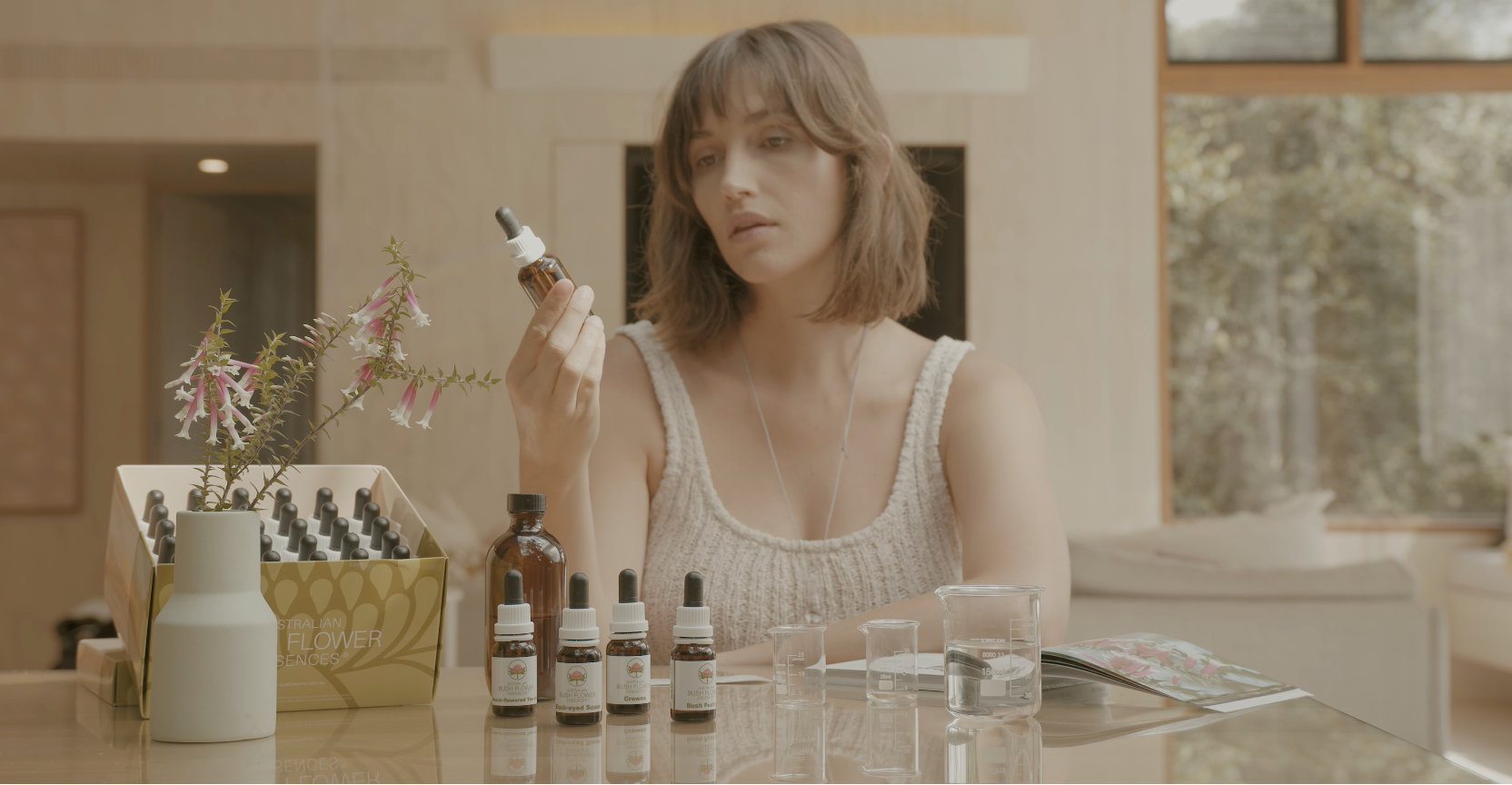 Woman holding a dropper bottle with various bottles and boxes on a table in a room with large windows.